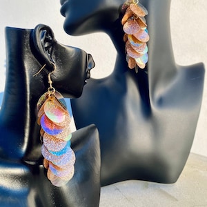 Könnte beinhalten: Hängende Ohrringe mit schimmernden, paillettenartigen Scheiben in Gold-, Rosa- und Blautönen. Die Ohrringe sind an goldfarbenen Haken befestigt. Die Ohrringe werden auf einem schwarzen Schaufensterpuppenkopf präsentiert.