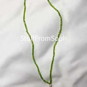 May include: A beaded necklace with varying shades of green beads. The necklace has a small gold-colored dinosaur pendant. The text "StuffFromSoph" is visible on the white background.