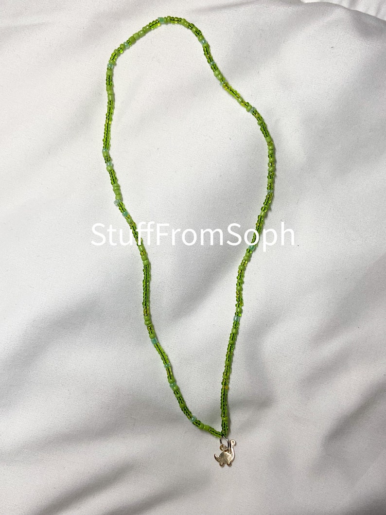 May include: A beaded necklace with varying shades of green beads. The necklace has a small gold-colored dinosaur pendant. The text "StuffFromSoph" is visible on the white background.