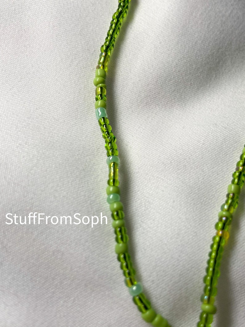 May include: Close-up of a beaded necklace with a variety of green beads. The beads are strung together to create a delicate and intricate design. The necklace is set against a white background. The text "StuffFromSoph" is visible.