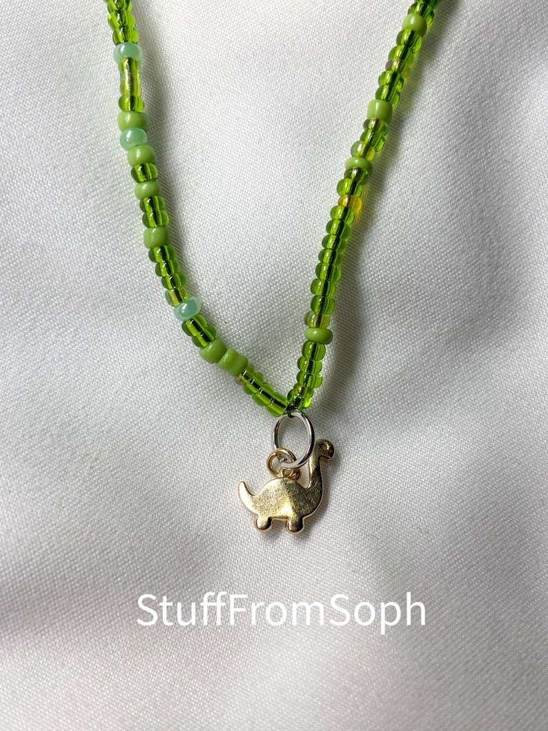 May include: A beaded necklace with a gold-colored dinosaur pendant. The necklace features a string of small, green beads in varying shades. The pendant is a stylized dinosaur shape. The text "StuffFromSoph" is visible in the lower part of the image.