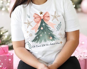 May include: White t-shirt with a Christmas tree design featuring a pink bow, gold stars, and the text "Oh Come Let Us Adore Him".