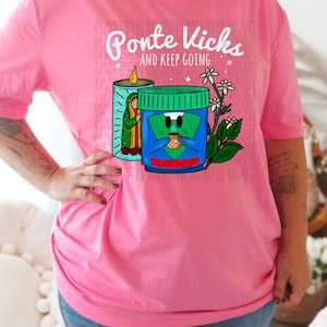 May include: Pink t-shirt with a graphic of a Vicks VapoRub container with a face and a candle. The text "Ponte Vicks - And Keep Going" is printed on the shirt.