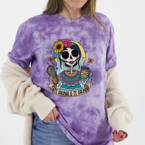 以下が含まれることがあります： 紫色のタイダイ染めのTシャツに、花柄のヘッドバンドをつけた骸骨がコーヒーカップとペストリーを持っているという漫画のイラストが描かれています。イラストの下には「¿Te calmas o te calmo? 」という文字が印刷されています。