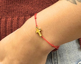 May include: A red string bracelet with a gold-colored cross charm. The bracelet is worn on a wrist, with the person's hand in a blue jean pocket. The background includes a rust-colored sweater and a tattoo on the arm.