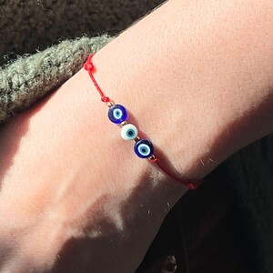 Pu&ograve; includere: Un braccialetto di filo rosso con tre perline a forma di occhio blu e bianco. Il braccialetto &egrave; indossato su un polso, con un maglione verde e pantaloni di velluto a coste marroni sullo sfondo.