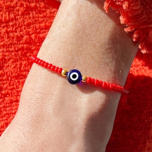 Puede incluir: Una pulsera delicada que presenta una cuenta central con forma de ojo turco en azul, blanco y negro, flanqueada por dos pequeñas cuentas metálicas doradas, ensartadas en una banda continua de pequeñas cuentas circulares de color rojo vivo.