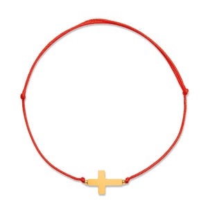 Puede incluir: Una pulsera con un dije de cruz dorada y un cordón rojo brillante. La cruz está centrada en el cordón y tiene un diseño sencillo. La pulsera se muestra sobre un fondo blanco.