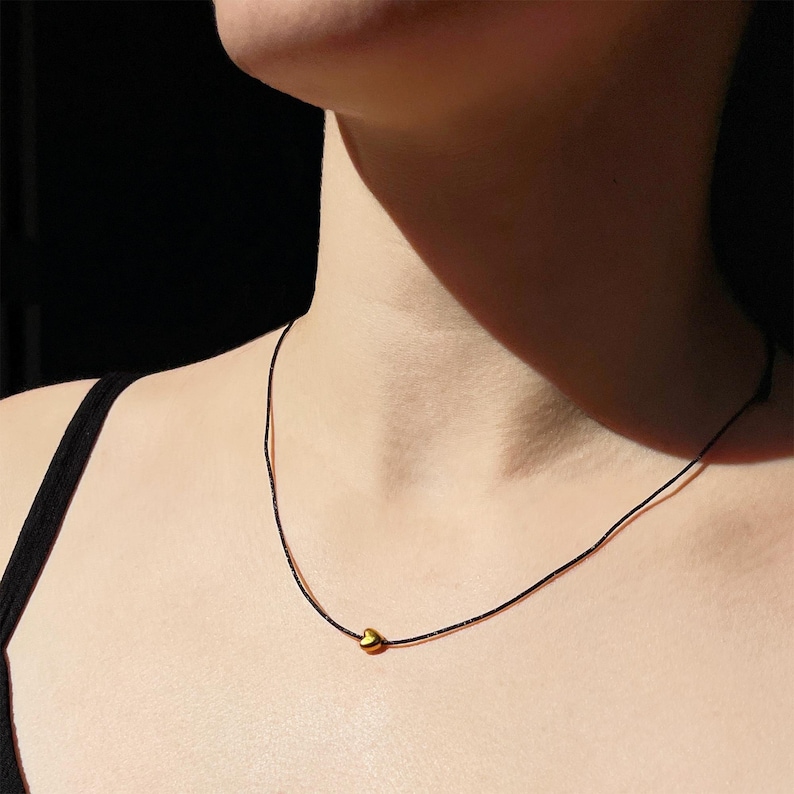 K&ouml;nnte beinhalten: Eine minimalistische Halskette mit einem kleinen, goldfarbenen Herzanh&auml;nger an einem d&uuml;nnen, schwarzen Band. Die Halskette liegt eng am Hals an und erzeugt einen schlichten und eleganten Look. Der dunkle Hintergrund hebt den Schmuck hervor.