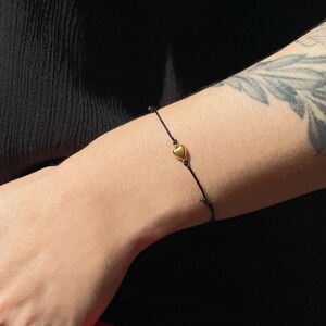 May include: A black string bracelet with a small, gold-colored heart charm. The bracelet is worn on a wrist, and the background is a dark, solid color. The heart charm is centered on the bracelet.