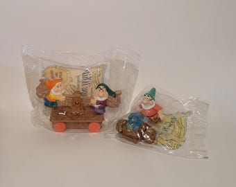 May include: Two sealed plastic bags containing Snow White and the Seven Dwarfs figurines. One bag has three dwarfs on a brown cart with orange wheels. The other bag has a dwarf with a blue object.