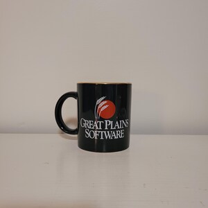 May include: A black ceramic mug featuring a circular red graphic with white wheat stalks and the white text Great Plains Software printed on the front. The mug has a curved handle on the left side and a smooth, glossy finish throughout the exterior.