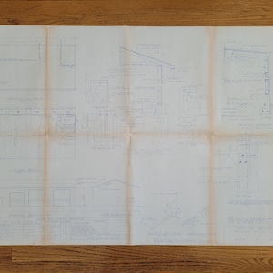 May include: A blueprint for a building with multiple sections, including a floor plan, side elevations, and details of the roof and windows. The blueprint is drawn in blue ink on white paper and is folded in several places.