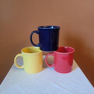 May include: A set of three ceramic mugs featuring a smooth, glossy finish. One mug is deep navy blue, one is bright yellow, and one is a vibrant coral red. Each mug has a rounded handle and a slightly flared rim, arranged in a stacked formation.