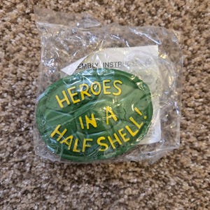 May include: A green, turtle-shaped item with the words "HEROES IN A HALF SHELL!" in yellow lettering. The item is in a clear plastic package, resting on a brown, textured surface.