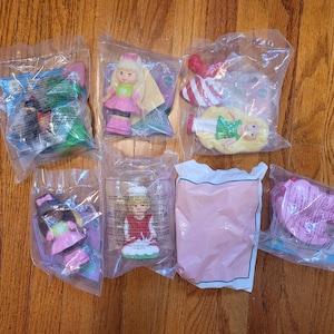 May include: Eight unopened packages of plastic dolls, each in a clear plastic bag. The dolls are dressed in various colorful outfits, including pink, green, and red. Some of the packages have a white background with text, while others have a clear background.