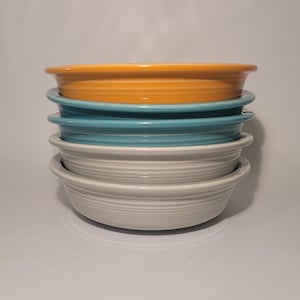 May include: A stack of four ceramic bowls in a variety of colors. The top bowl is orange, followed by a teal bowl, and two white bowls at the bottom. Each bowl has a ridged design and a rounded rim.
