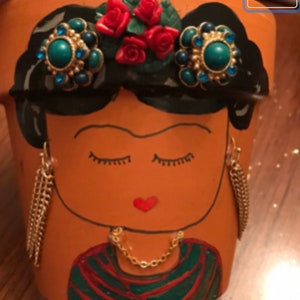 May include: Terracotta flower pot featuring a painted face with closed eyes, red heart-shaped lips, and gold chain earrings. The pot is adorned with a black painted hairstyle, red rose accents, and turquoise and gold embellishments.