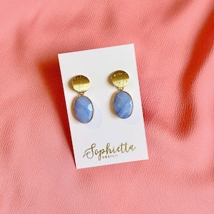 Könnte beinhalten: Ein Paar goldfarbene Ohrringe mit ovalen, hellblauen Edelsteinen. Die Ohrringe werden auf einer weißen Karte mit dem Markennamen "Sophietta Design" in Schreibschrift präsentiert. Der Hintergrund ist ein rosa Stoff.