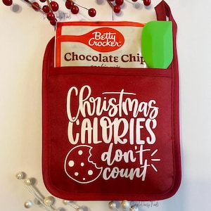 May include: Red oven mitt with white text that reads "Christmas Calories don't count". A white cookie with a bite taken out of it is next to the text. A green spatula and a bag of Betty Crocker chocolate chip cookie mix are in the pocket of the oven mitt.