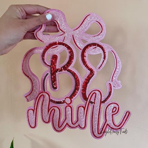 May include: Pink and red sequin patch with the text "Be Mine" in a cursive font.