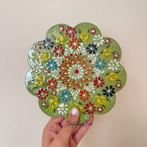 May include: A green ceramic plate with a floral design. The plate is decorated with white, red, yellow, blue, and black flowers. The plate is hand-painted and has a unique, whimsical design.
