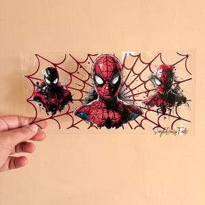 Puede incluir: Una pegatina rectangular transparente con tres ilustraciones de Spider-Man. La imagen central muestra un busto detallado de Spider-Man rojo y negro, flanqueado por otras dos figuras de Spider-Man. Un dise&ntilde;o de telara&ntilde;a roja enmarca las im&aacute;genes. El texto "Simple Daisy Finds" es visible.