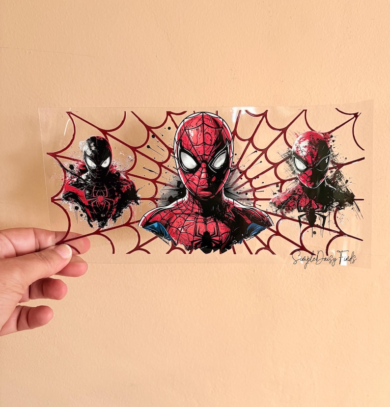 Puede incluir: Una pegatina rectangular transparente con tres ilustraciones de Spider-Man. La imagen central muestra un busto detallado de Spider-Man rojo y negro, flanqueado por otras dos figuras de Spider-Man. Un dise&ntilde;o de telara&ntilde;a roja enmarca las im&aacute;genes. El texto "Simple Daisy Finds" es visible.