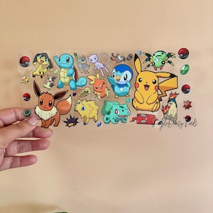 May include: A clear, rectangular sticker sheet featuring various colorful cartoon characters, including Pikachu, Squirtle, and Eevee. The sheet is held by a hand against a neutral background, with additional decorative elements like Pok&eacute; Balls and gems.