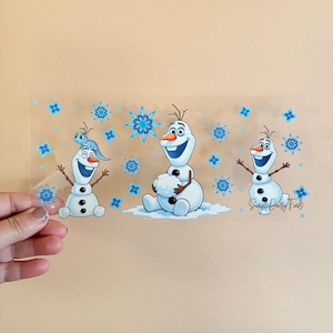 May include: A clear sticker featuring three cartoon snowmen with smiling faces and stick arms. The snowmen are white with black button eyes and carrot noses. Blue snowflakes and the text "Supple Daisy Finds" are also present.
