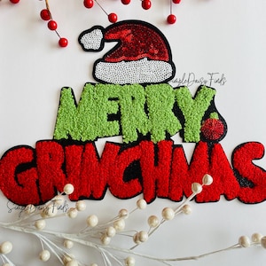 May include: A festive Christmas decoration with the words "MERRY GRINCHMAS" in green and red, with a Santa hat. The text is textured and outlined in black. Decorative berries and branches are in the background.