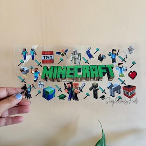 May include: A clear sheet featuring the word "Minecraft" in green block letters, surrounded by pixelated characters, swords, and other game-related icons. The sheet is held by a hand against a beige background.
