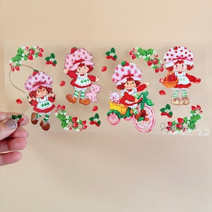 May include: A sheet of stickers featuring four Strawberry Shortcake characters in various poses. The characters are wearing red and white outfits and strawberry-themed hats. The stickers include strawberries and green leaves.
