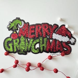 May include: A sequined Christmas decoration with the words "MERRY GRINCHMAS" in red and green. The design includes a Grinch-themed Christmas tree and Santa hat. A string of red glittery berries is in the foreground.