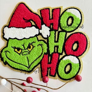 May include: A Christmas-themed applique featuring the Grinch wearing a Santa hat, with the words "HO HO HO". The design is primarily green, red, and white, with a gold glitter border. Decorative berries and branches are in the foreground.