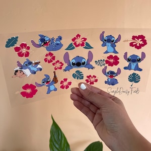 May include: A clear sheet featuring multiple blue cartoon character illustrations with large ears, surrounded by pink hibiscus flowers and green leaves. The sheet is held by a hand with white nail polish. The text "SimpleDaisyFinds" is visible.