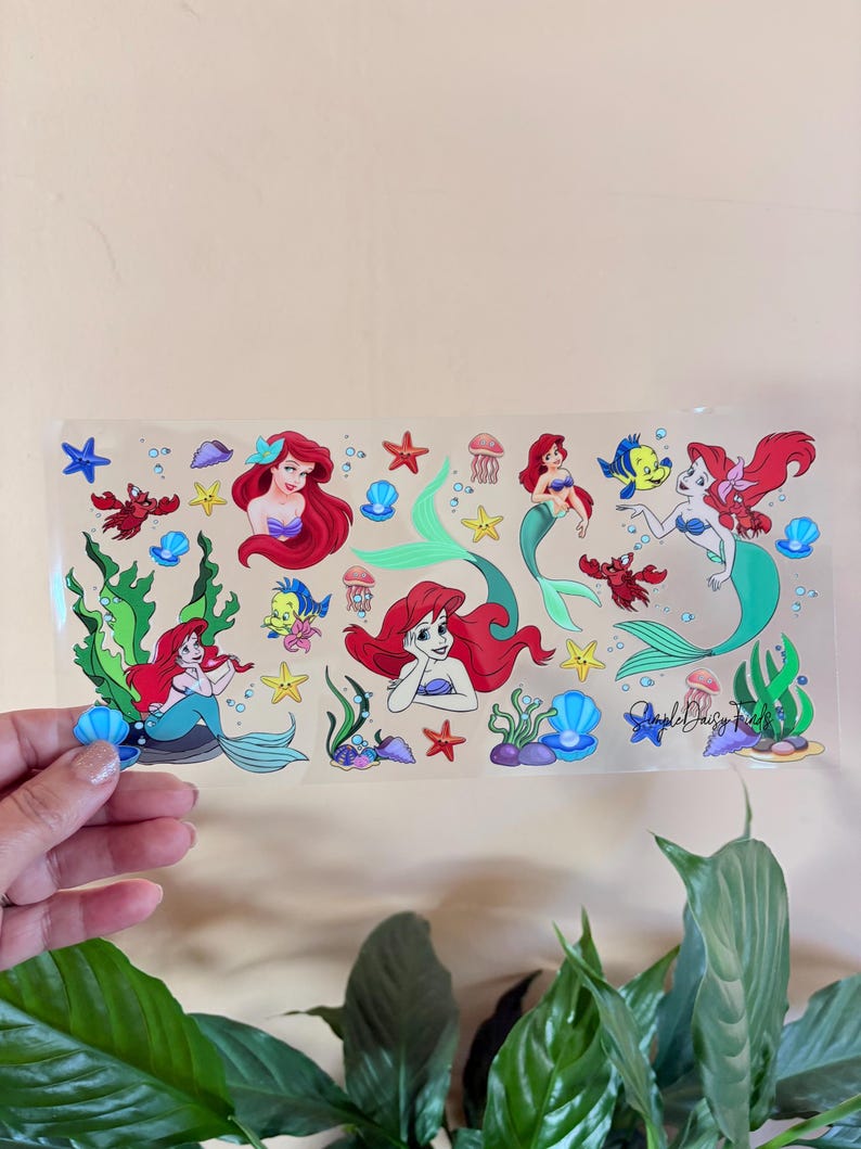 May include: A clear sticker sheet featuring illustrations of a mermaid with red hair, sea creatures, and aquatic plants. The sticker sheet includes the text "Simple Daisy Finds".