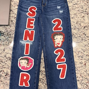 Op de afbeelding: Donkerblauwe spijkerbroek met rode glitterletters die SENIOR op de linkerpijp en 27 op de rechterpijp vormen. Het ontwerp bevat twee grafische patches van het stripfiguur Betty Boop. De spijkerbroek heeft een versleten afwerking.