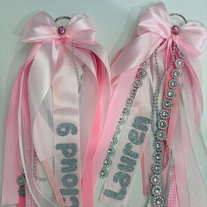 May include: Two decorative pink satin ribbon corsages featuring silver glitter lettering that reads Cloud 9 and Lauren. Each corsage is topped with a pink satin bow, a central pink rhinestone accent, and hanging silver beaded chains and floral charms.