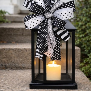 May include: Black metal lantern with a lit candle inside, topped with a large bow. The bow features black and white polka dot, striped, and patterned ribbons. The lantern is sitting on a stone step.