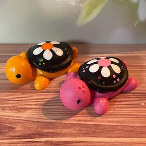May include: Two ceramic turtle figurines. One is orange with a black shell decorated with a white and orange flower. The other is pink with a black shell decorated with a white and pink flower. The turtles are on a wooden surface.