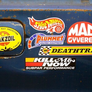 May include: A blue car with various stickers on the back. The stickers include "Hot Wheels", "Plummer Racing Equipment", "Mac Gyvered", "Death Trap", "Kill M& Now Subpar Performance", and "Perpetual Rust Protection Leakzoil If It Leaks Oil It Has Oil".
