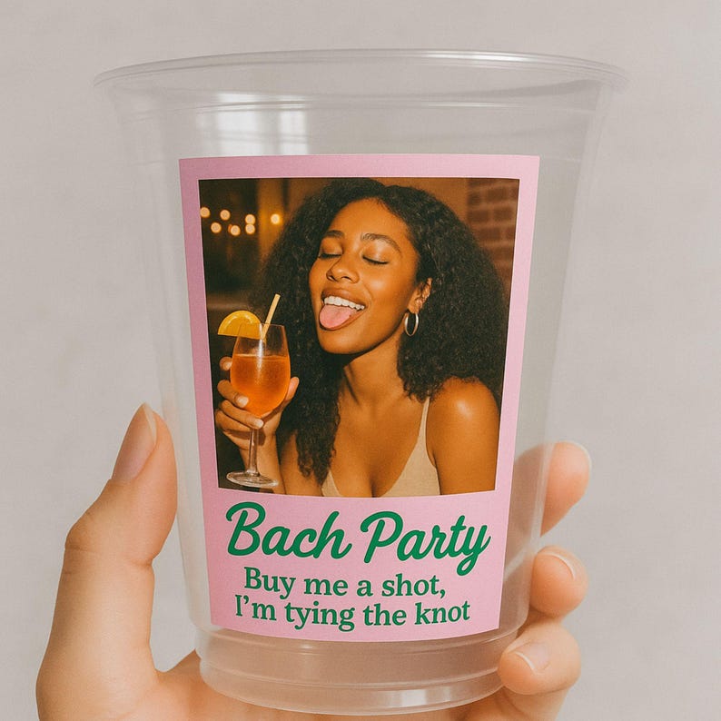 May include: Clear plastic cup with a pink label featuring a photo of a woman holding a cocktail. The label reads "Bach Party" and "Buy me a shot, I'm tying the knot."