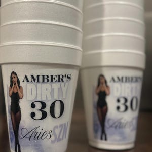 May include: White foam cups with a black and white design featuring a woman in a black outfit and the text "Amber's Dirty 30 Aries SZN".