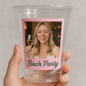 May include: Clear plastic cup with a pink label featuring a photo of a smiling woman holding a wine glass. The label has the words "Bach Party" in green script. The cup is held by a hand.
