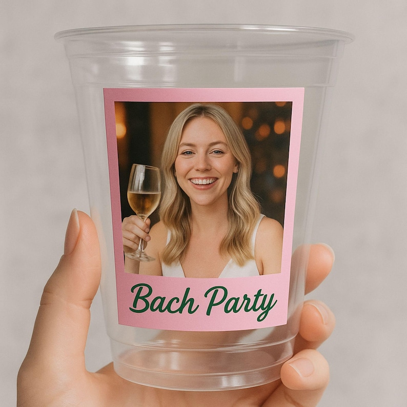 May include: Clear plastic cup with a pink label featuring a photo of a smiling woman holding a wine glass. The label has the words "Bach Party" in green script. The cup is held by a hand.