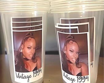 May include: Two clear plastic cups with lids. Each cup has a label featuring a photo of a woman with the text "Vintage 1994". The cups are on a light brown surface.