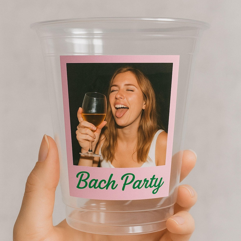 May include: Clear plastic cup with a pink label featuring a photo of a person holding a wine glass and sticking out their tongue. The words "Bach Party" are printed in green on the label. The cup is held by a hand.