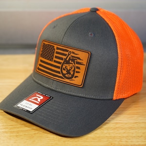 May include: A gray and orange trucker hat with a brown leather patch featuring an American flag and a deer silhouette. The patch has the text "Flexfit" on it.
