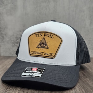 May include: White and black trucker hat with a brown patch that says "Tin Foil Conspiracy Realist" and features an all-seeing eye graphic. The hat has a black mesh back and an adjustable strap with a black label that says "Richardson 112 Trucker".